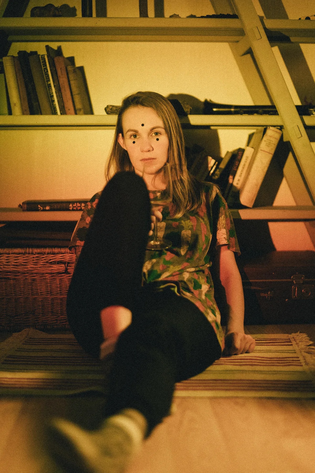 A woman seated beside bookshelves in warm indoor light.