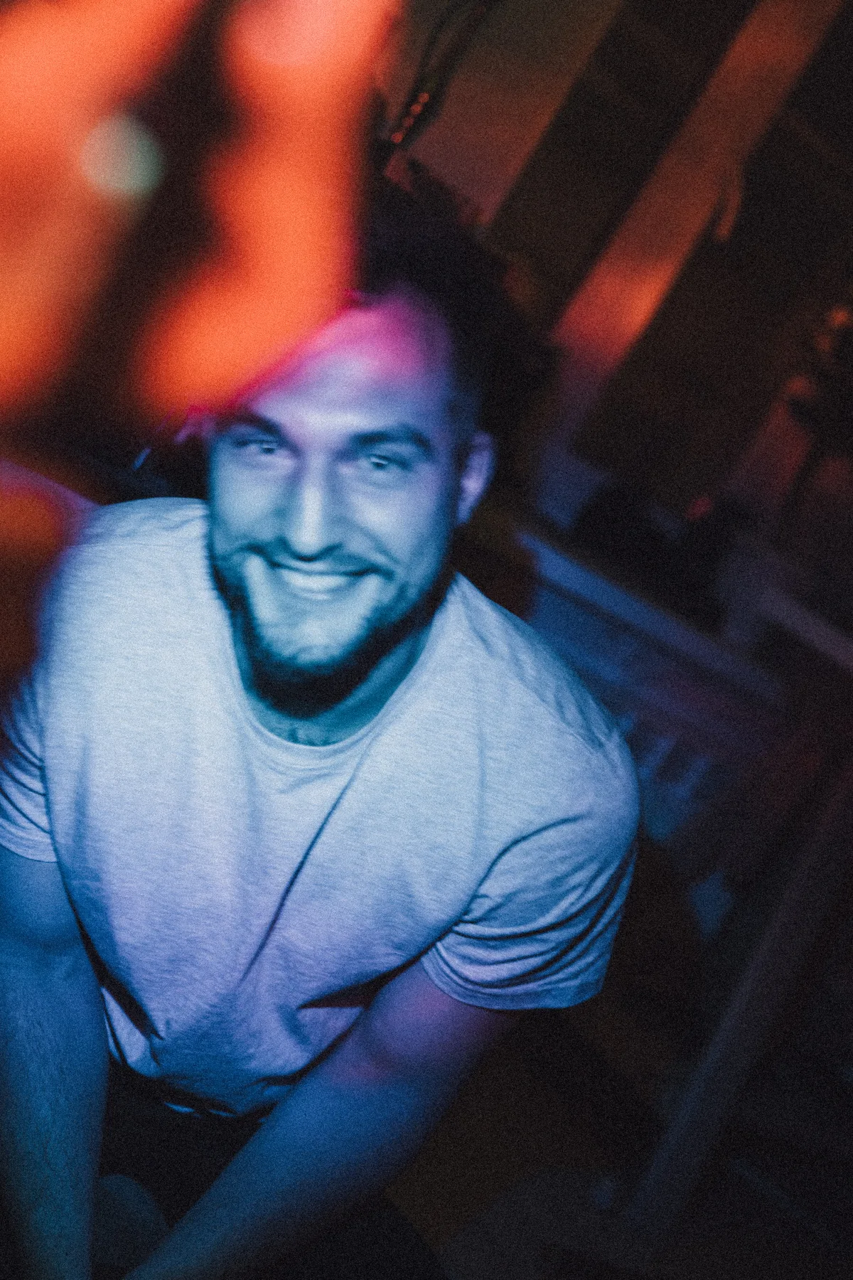 A smiling man leaning towards the camera beneath blue and orange light.