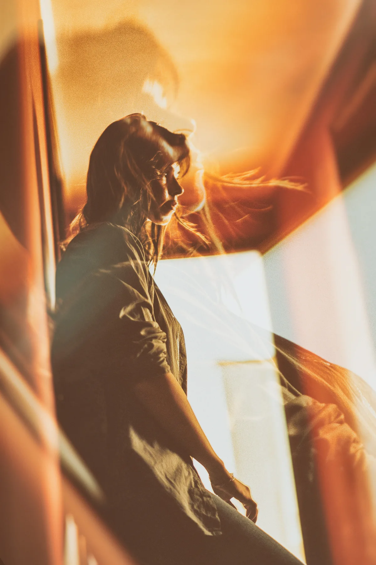 A woman standing beside a bright window amid warm reflections.