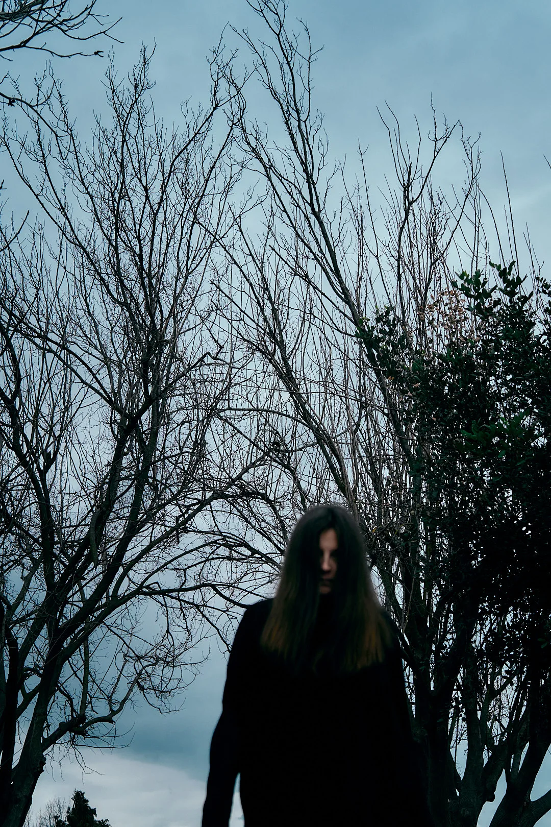 A dark-haired woman standing beneath bare branches against a blue-grey sky.