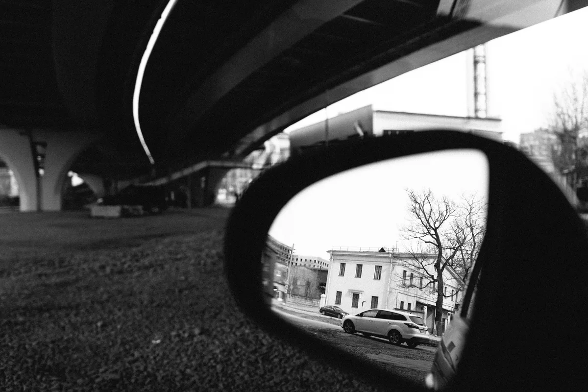 Black-and-white view of a building reflected in a car mirror beneath an overpass.