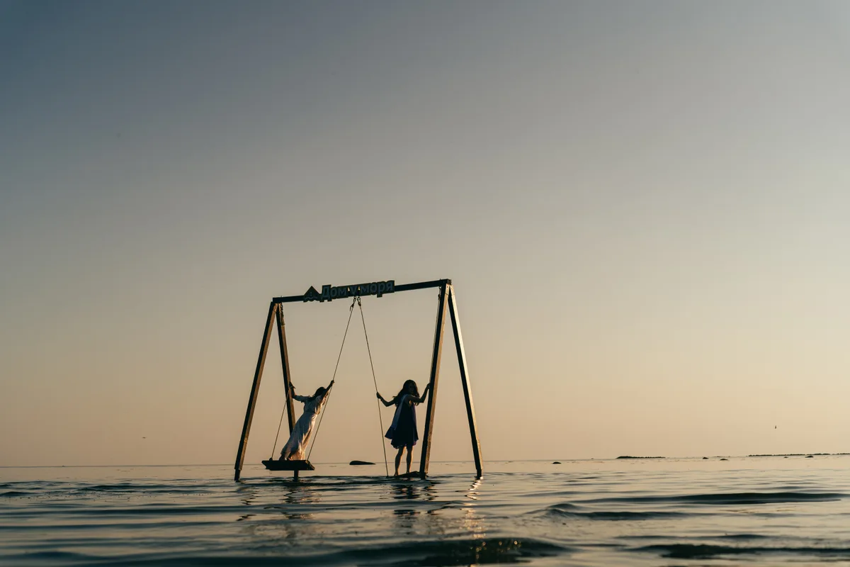 People on swings standing in shallow water beneath an open sky.