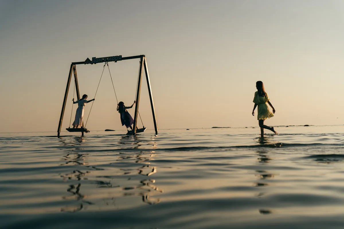 A child walking through shallow water towards people on swings.