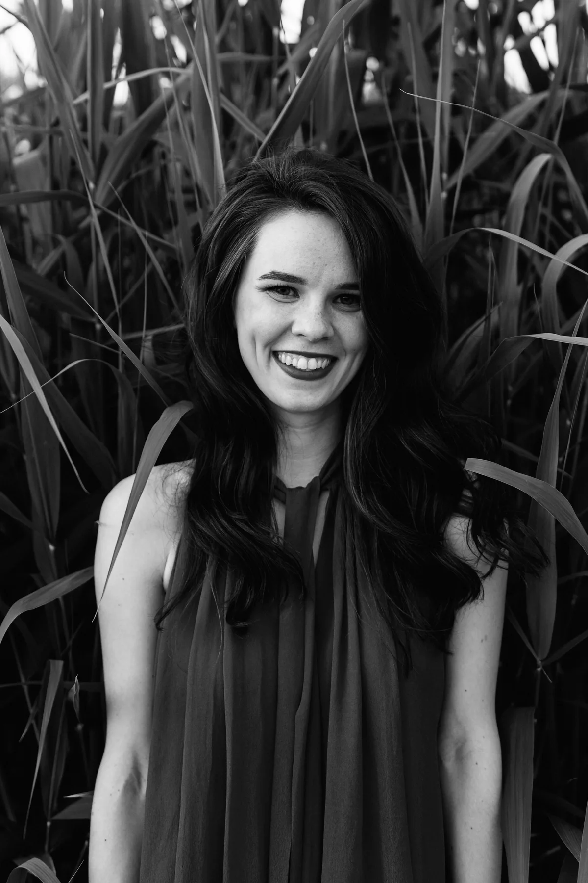 Black-and-white portrait of a smiling woman against tall leaves.