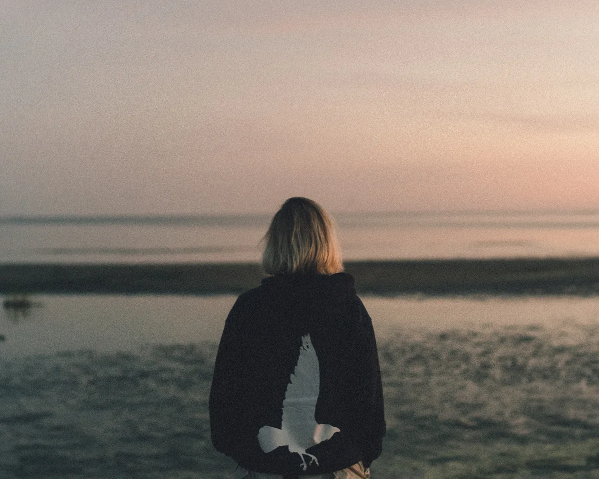 A person seen from behind on a beach beneath a pale pink sky.